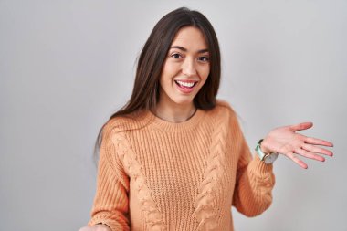 Young brunette woman standing over white background smiling cheerful with open arms as friendly welcome, positive and confident greetings 