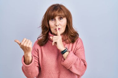 Orta yaşlı İspanyol bir kadın izole edilmiş arka planda duruyor. Parmakları dudaklarında, elleri yana dönük bir şekilde sessiz olmak istiyor. sessizlik ve gizli konsept. 
