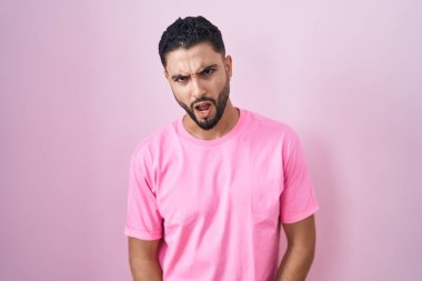 Hispanic young man standing over pink background in shock face, looking skeptical and sarcastic, surprised with open mouth 