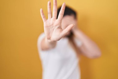 Young brunette woman standing over yellow background covering eyes with hands and doing stop gesture with sad and fear expression. embarrassed and negative concept. 