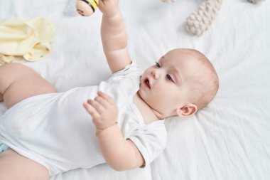 Beyaz bebek yatakta uzanmış, yatak odasında Maraca oyuncağı tutuyor.