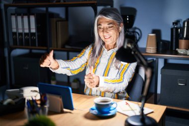 Gri saçlı orta yaşlı bir kadın gece ofiste çalışıyor. Elleriyle olumlu jest yapmayı onaylıyor. Gülümsüyor ve başarı için seviniyor. kazanan jest. 