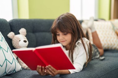 Adorable hispanic girl reading book lying on sofa at home