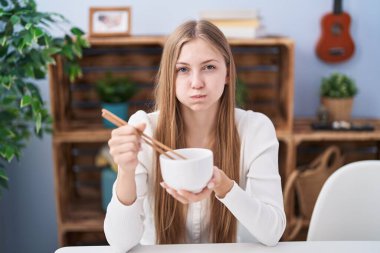 Young caucasian woman eating asian food using chopsticks puffing cheeks with funny face. mouth inflated with air, catching air. 