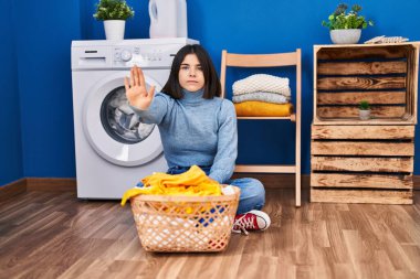 Young hispanic woman at laundry room with open hand doing stop sign with serious and confident expression, defense gesture 
