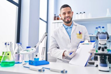 Hispanic man working at scientist laboratory holding blank clipboard clueless and confused expression. doubt concept. 