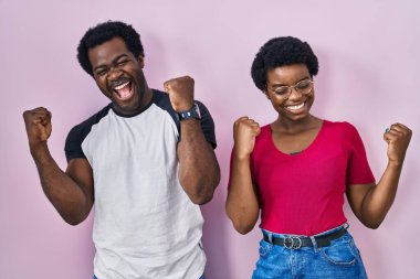 Genç Afro-Amerikan çifti pembe arka planda durup şaşkınlık ve şaşkınlık içinde kollarını kaldırarak ve gözlerini kapatarak başarıyı kutluyor. Kazanan konsept. 