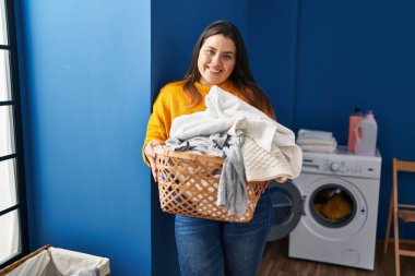 Young beautiful plus size woman smiling confident holding basket with clothes at laundry room