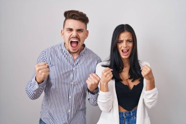 Young hispanic couple standing over white background angry and mad raising fists frustrated and furious while shouting with anger. rage and aggressive concept. 