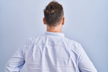 Orta yaşlı beyaz bir adam mavi arka planda dikiliyor geriye bakıyor kolları vücudunda. 