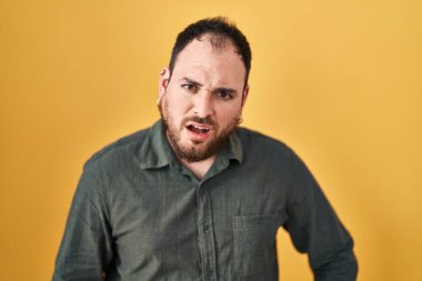 Plus size hispanic man with beard standing over yellow background in shock face, looking skeptical and sarcastic, surprised with open mouth 