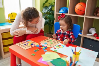 Teacher and toddler playing with maths puzzle game sitting on table at kindergarten