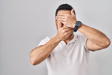 Beyaz arka plan üzerine günlük kıyafetler giyen, ağzı ve gözlerini kapayan, şaşırmış ve şok olmuş genç İspanyol bir adam. duygu gizleme 