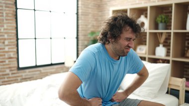 Young hispanic man suffering for stomach ache sitting on bed at bedroom