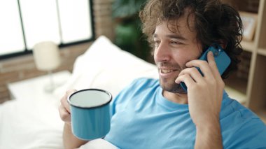 Genç İspanyol adam akıllı telefonda konuşuyor. Yatak odasında kahve içiyor.