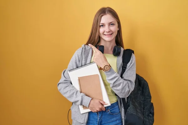 Young caucasian woman wearing student backpack and holding books with a big smile on face, pointing with hand finger to the side looking at the camera.