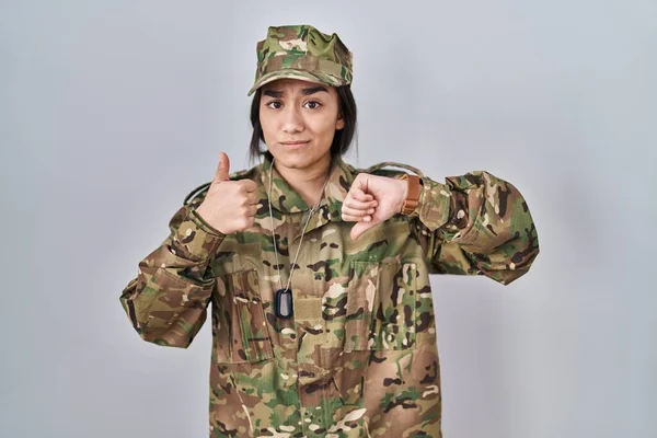 Young south asian woman wearing camouflage army uniform doing thumbs up and down, disagreement and agreement expression. crazy conflict