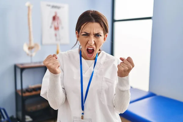 Young brunette woman working at pain recovery clinic angry and mad raising fists frustrated and furious while shouting with anger. rage and aggressive concept.