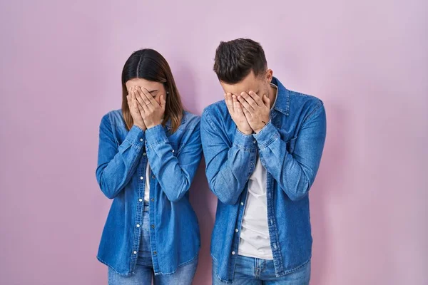 Young hispanic couple standing over pink background with sad expression covering face with hands while crying. depression concept.