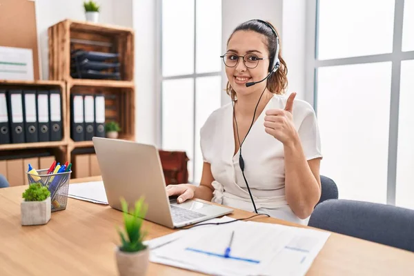 Young hispanic woman wearing call center agent headset at the office smiling happy and positive, thumb up doing excellent and approval sign