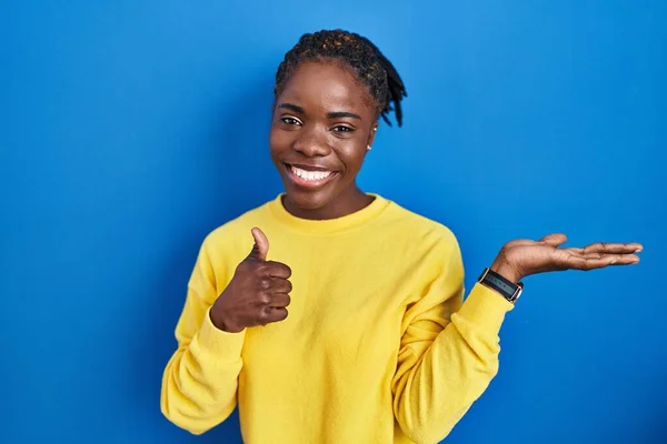 Beautiful black woman standing over blue background showing palm hand and doing ok gesture with thumbs up, smiling happy and cheerful
