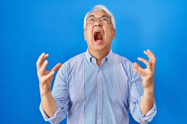 Hispanic senior man wearing glasses crazy and mad shouting and yelling with aggressive expression and arms raised. frustration concept.