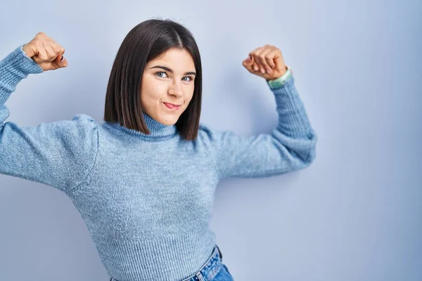 Young hispanic woman standing over blue background showing arms muscles smiling proud. fitness concept. 