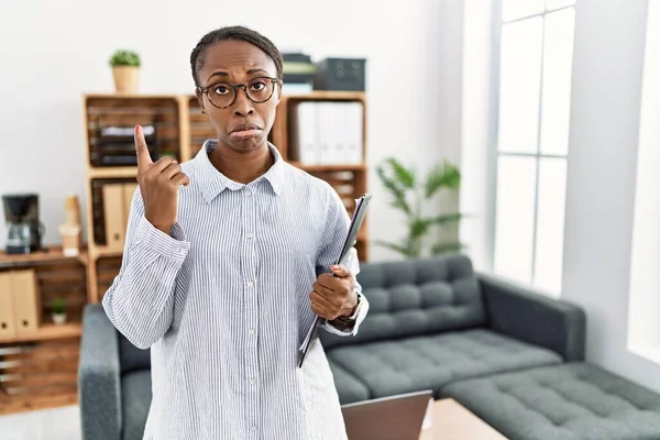African woman working at psychology clinic pointing up looking sad and upset, indicating direction with fingers, unhappy and depressed.