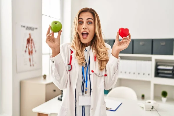 Young Blonde Doctor Woman Holding Heart Green Apple Celebrating Crazy — 图库照片