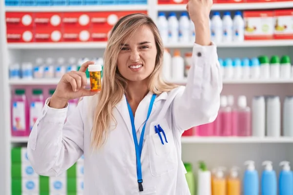 Young blonde woman working at pharmacy drugstore holding pills annoyed and frustrated shouting with anger, yelling crazy with anger and hand raised