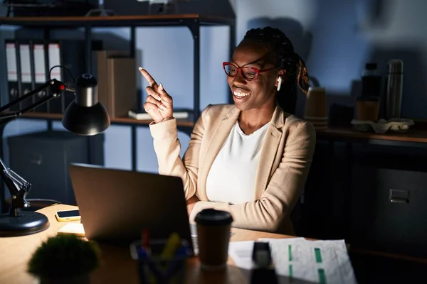 Beautiful black woman working at the office at night with a big smile on face, pointing with hand and finger to the side looking at the camera.