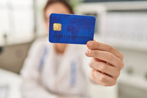 Young hispanic woman wearing doctor uniform holding credit card at clinic