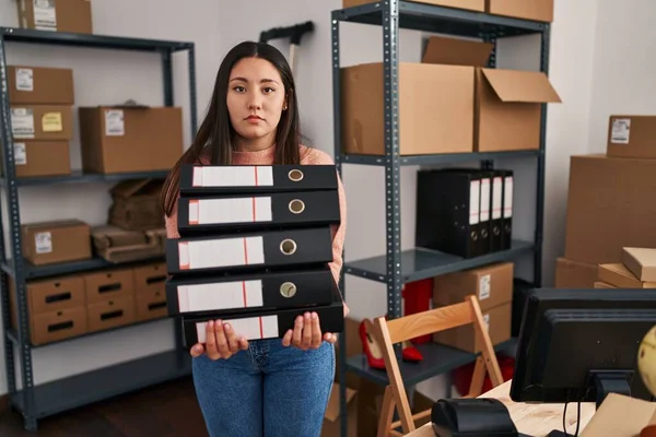 Young latin woman working at small business ecommerce holding folders relaxed with serious expression on face. simple and natural looking at the camera.