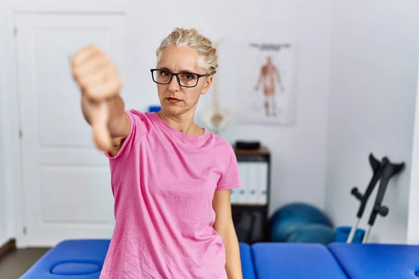 Middle age blonde woman at pain recovery clinic looking unhappy and angry showing rejection and negative with thumbs down gesture. bad expression.