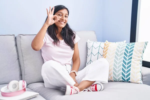 Young hispanic woman sitting on the sofa at home smiling positive doing ok sign with hand and fingers. successful expression.