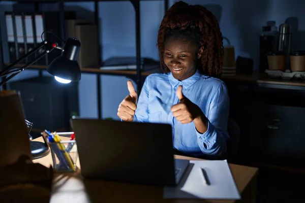 African woman working at the office at night approving doing positive gesture with hand, thumbs up smiling and happy for success. winner gesture.