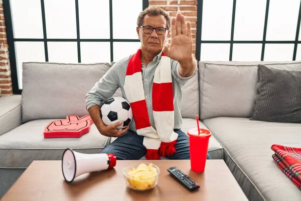 Senior man watching football holding ball supporting team with open hand doing stop sign with serious and confident expression, defense gesture