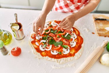Young beautiful hispanic woman putting onion on pizza at the kitchen