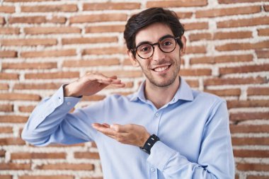 Young hispanic man standing over brick wall background gesturing with hands showing big and large size sign, measure symbol. smiling looking at the camera. measuring concept. 