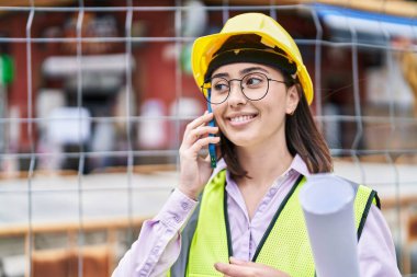 Young hispanic woman architect talking on the smartphone holding blueprints at street
