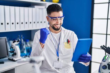Young handsome man working at scientist laboratory doing online call screaming proud, celebrating victory and success very excited with raised arm 
