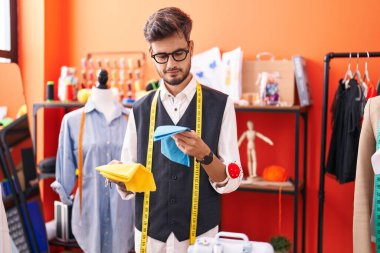 Young hispanic man tailor smiling confident holding cloths at atelier