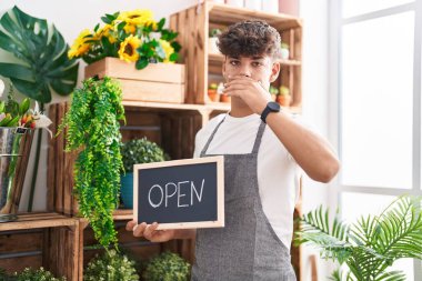 Hispanic teenager working at florist holding open sign covering mouth with hand, shocked and afraid for mistake. surprised expression 