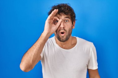Mavi arka planda dikilen Latin asıllı genç adam şaşırmış yüz ifadesiyle şaşırmış, parmaklarının arasından bakıyor. İnançsız ifade. 