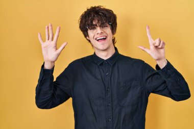 Sarı arka planda gözlük takan genç adam yedinci parmaklarını gösterip gülümserken kendinden emin ve mutlu görünüyor.. 