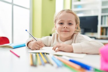 Adorable blonde girl student writing on notebook sitting on table at classroom