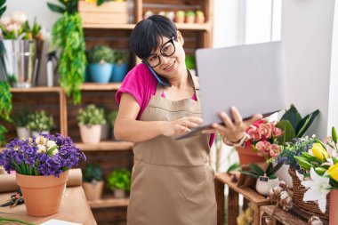 Middle age chinese woman florist talking on smartphone using laptop at flower shop