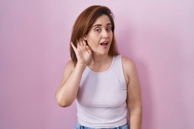 Brunette woman standing over pink background smiling with hand over ear listening an hearing to rumor or gossip. deafness concept. 
