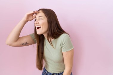Beautiful brunette woman standing over pink background very happy and smiling looking far away with hand over head. searching concept. 