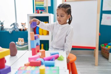 Adorable hispanic boy playing with construction blocks standing at kindergarten